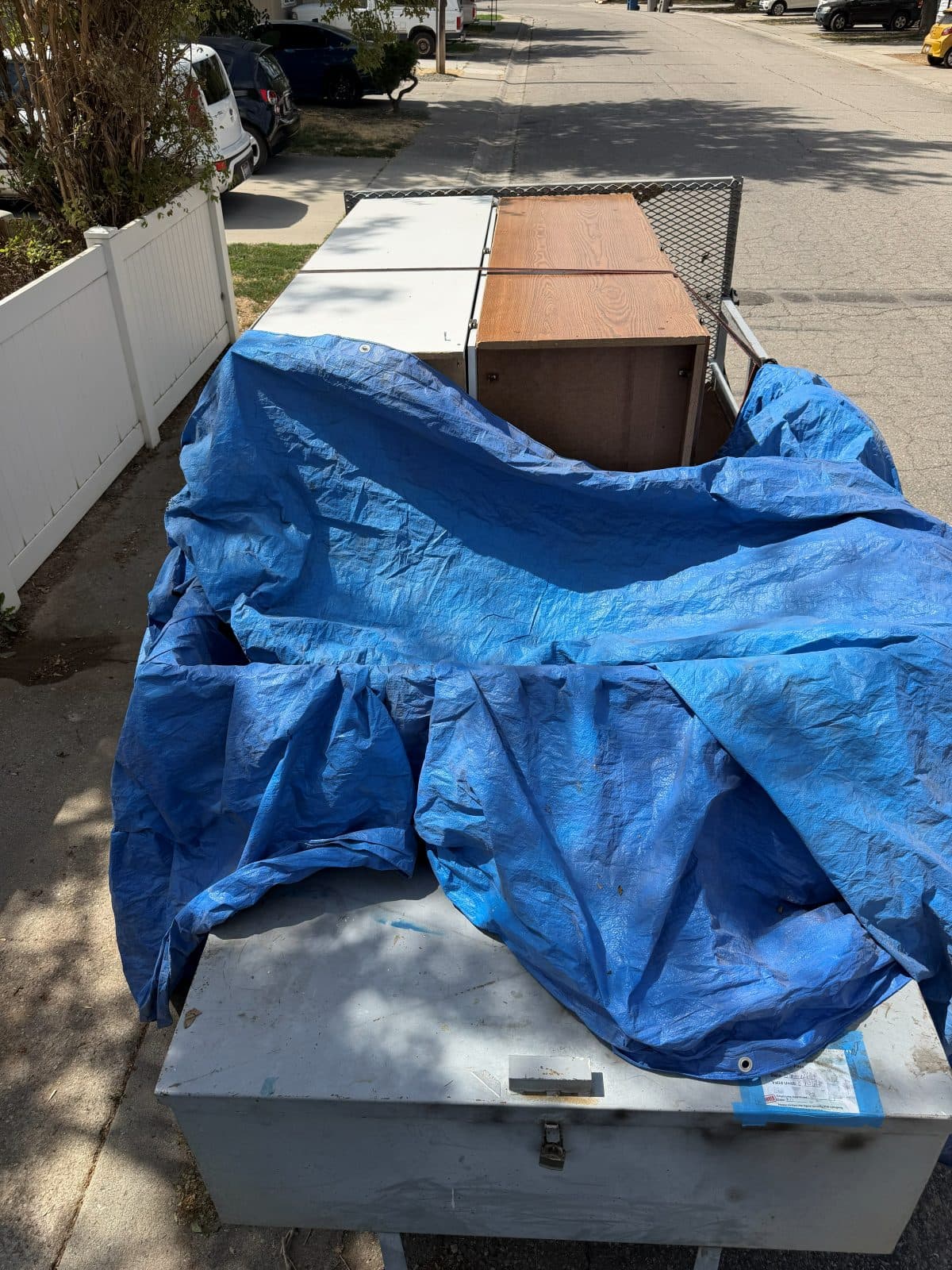 Trailer loaded with a wooden dresser and cabinet panels, being covered with a tarp on a residential street